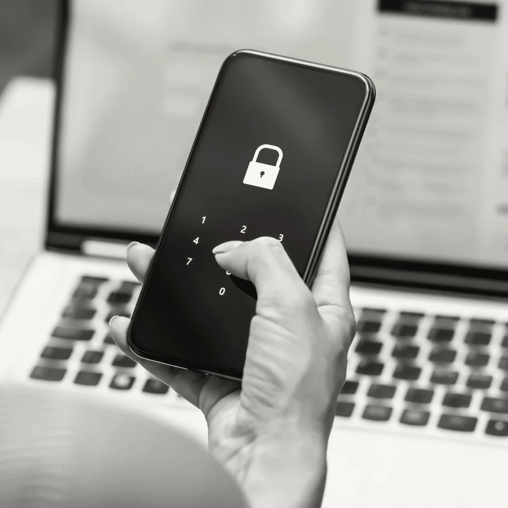 A hand entering a passcode on a smartphone with a lock icon on the screen, in front of a laptop.