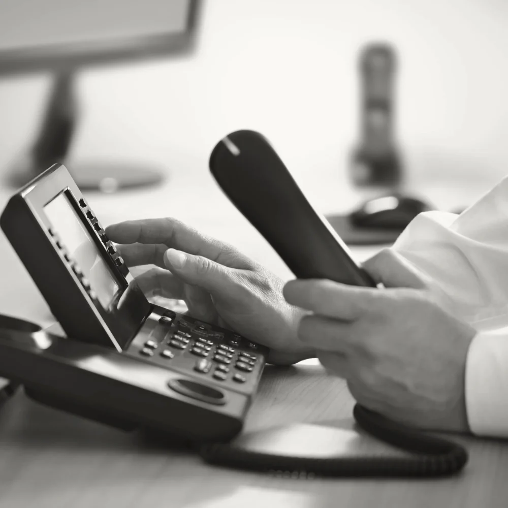 A close-up, black and white photograph of a person's hands using a desk phone.