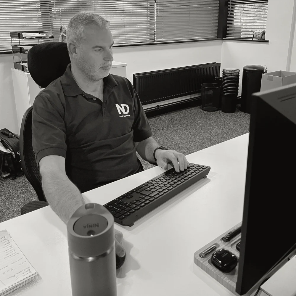 Male IT specialist wearing a Net-Defence branded polo shirt and working at a desk, typing on a keyboard while looking at a monitor.
